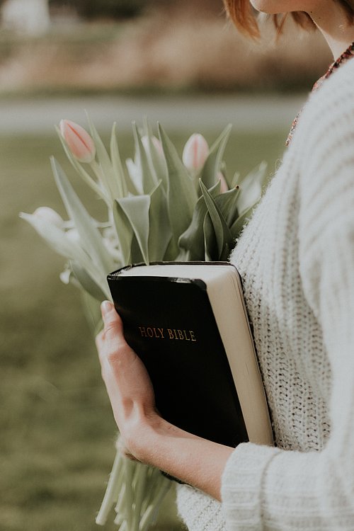 junge Frau mit Strickpullover hält eine Bibel in der linken und ein Strauß Tulpen in der rechten Hand.