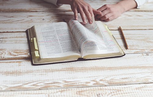 geöffnete Bibel auf weiß getünchtem Holz, ein Bleistift liegt rechts daneben, eine junge Frau blättert mit ihren Händen in der Bibel