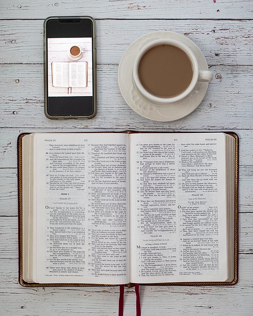geöffnete Bibel liegt auf weißen Holzplanken, rechts oben drüber steht eine cremefarbene Tasse mit Kaffee, links über der Bibel liegt ein Handy mit dem Bild der Bibel und der Tasse Kaffee